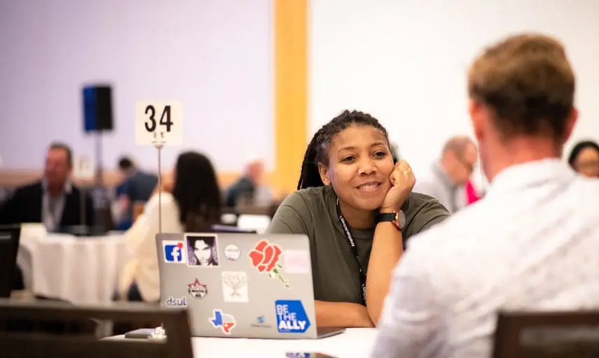 An African American woman sitting and leaning her cheek on her hand while sitting at a table slighting smiling.