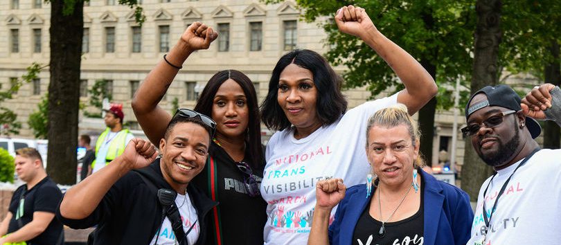 A group of four BIPOC holding up their closed fist in air, supporting trans rights.